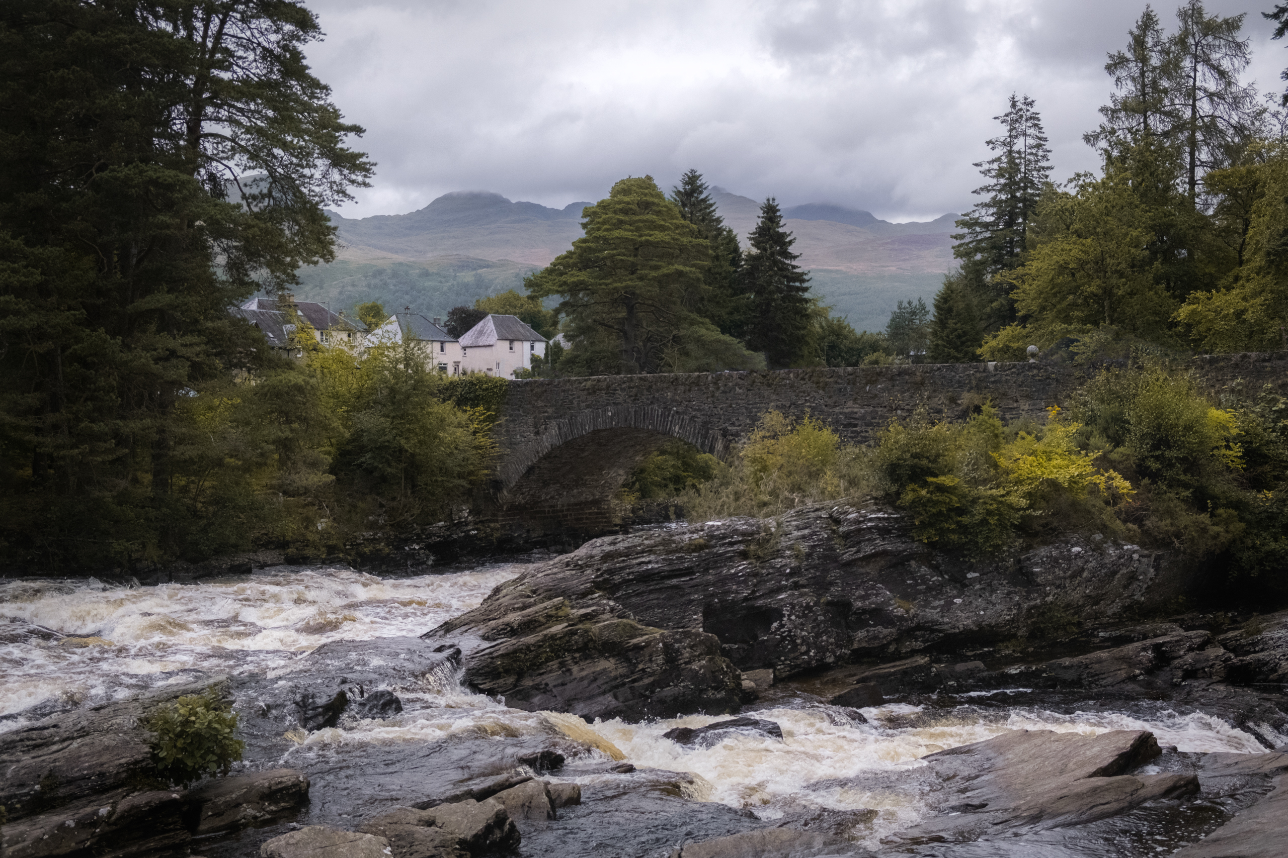 A fast flowing river in the scottish highlands with a bridge and houses in teh background